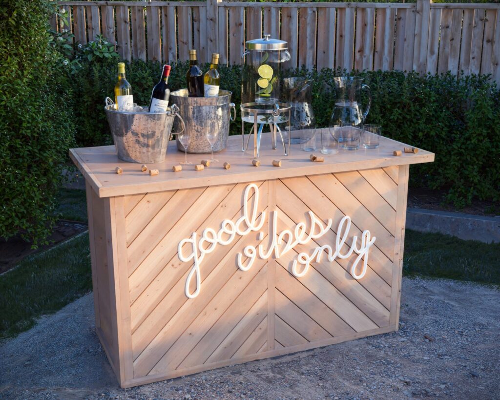 A beautifully crafted collapsible coffee bar with a wooden chevron pattern, featuring 'good vibes only' in white lettering, set up for an outdoor party with wine bottles, ice buckets, and glassware.