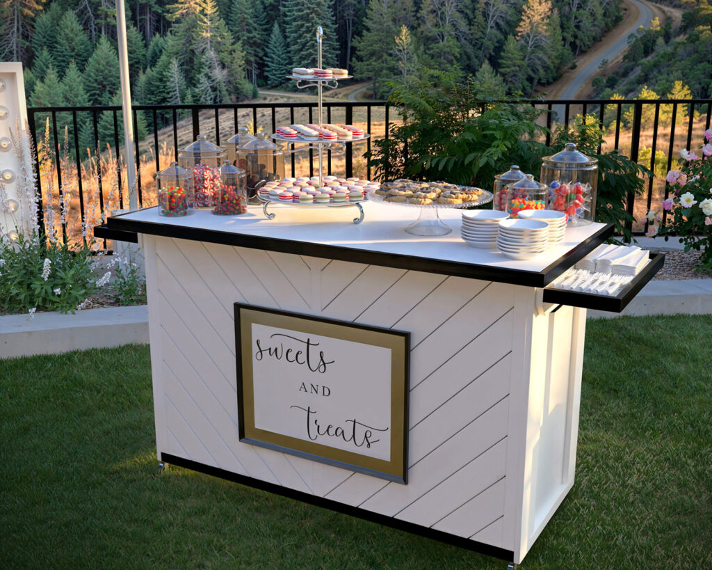 Elegant outdoor prom event with a folding event bar displaying desserts, surrounded by beautifully decorated tables, illuminated ‘PROM’ letters, and scenic mountain views.