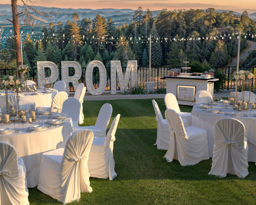 Elegant outdoor prom event with a folding event bar displaying desserts, surrounded by beautifully decorated tables, illuminated ‘PROM’ letters, and scenic mountain views.