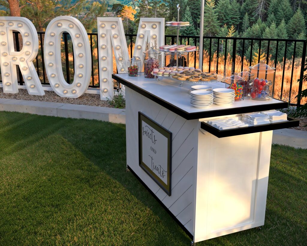 Elegant outdoor prom event with a folding event bar displaying desserts, surrounded by beautifully decorated tables, illuminated ‘PROM’ letters, and scenic mountain views.